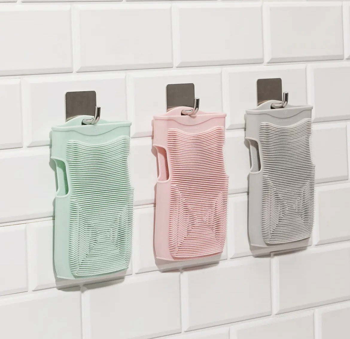 Three silicone body scrubbers in green, pink, and gray on a tiled wall.