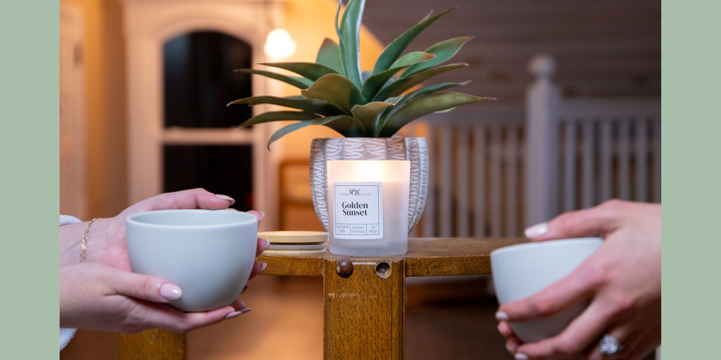 two people relaxing with a cup of tea with a Golden Sunset wooden wick candle between them, with a plant on the table