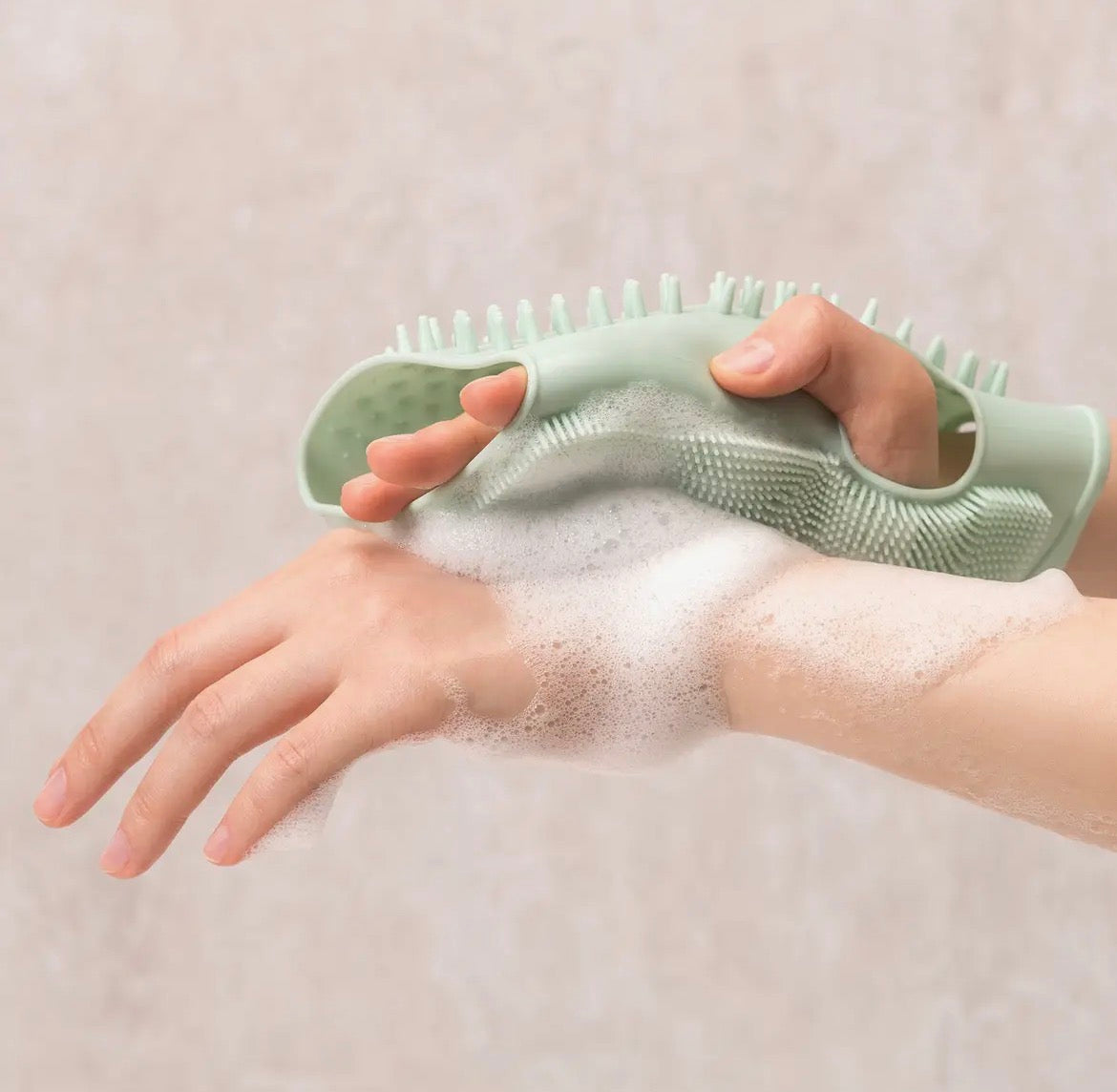 Person using a green scrub brush on a soapy hand against a beige background