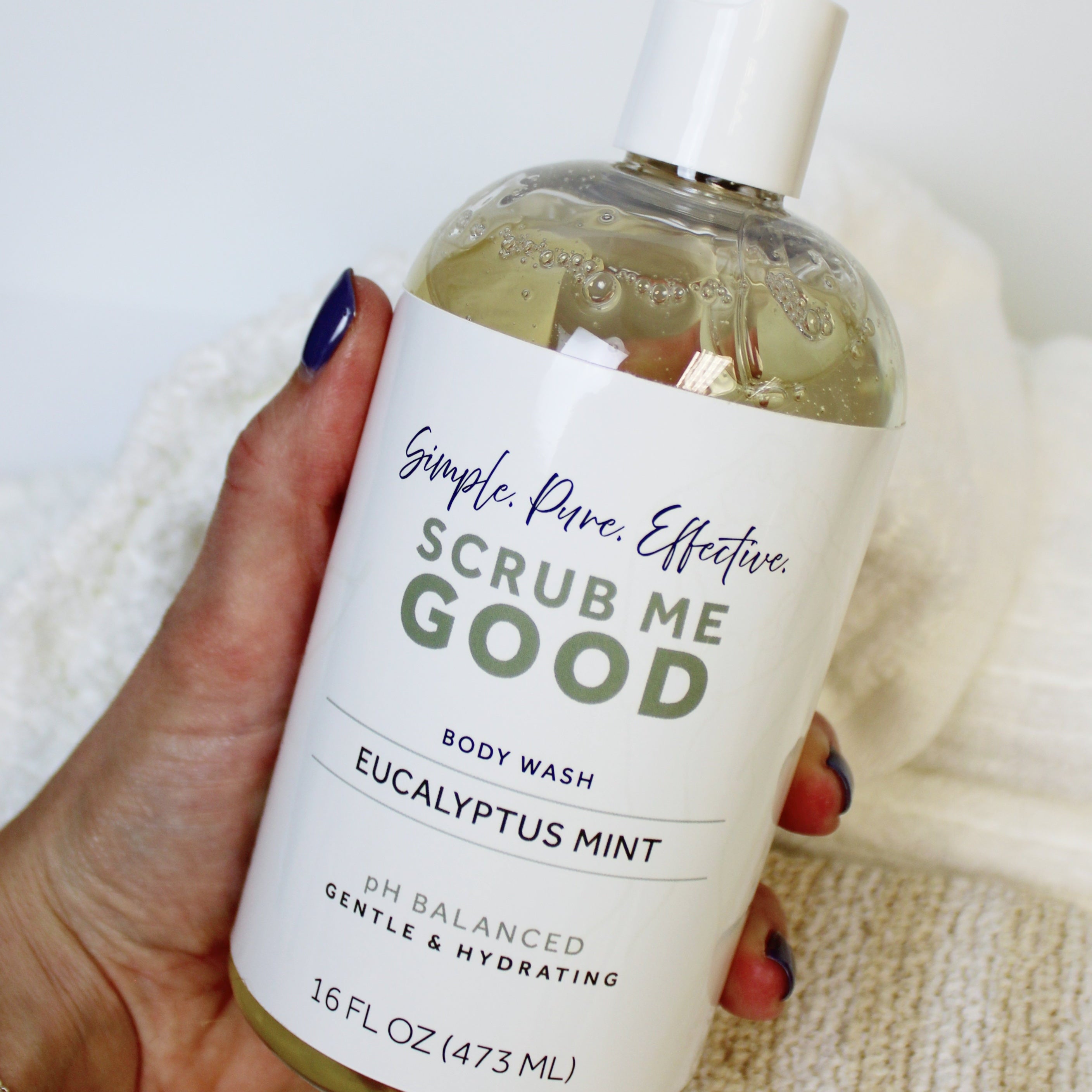 Hand holding a bottle of 'Scrub Me Good' Eucalyptus Mint body wash against a neutral background.
