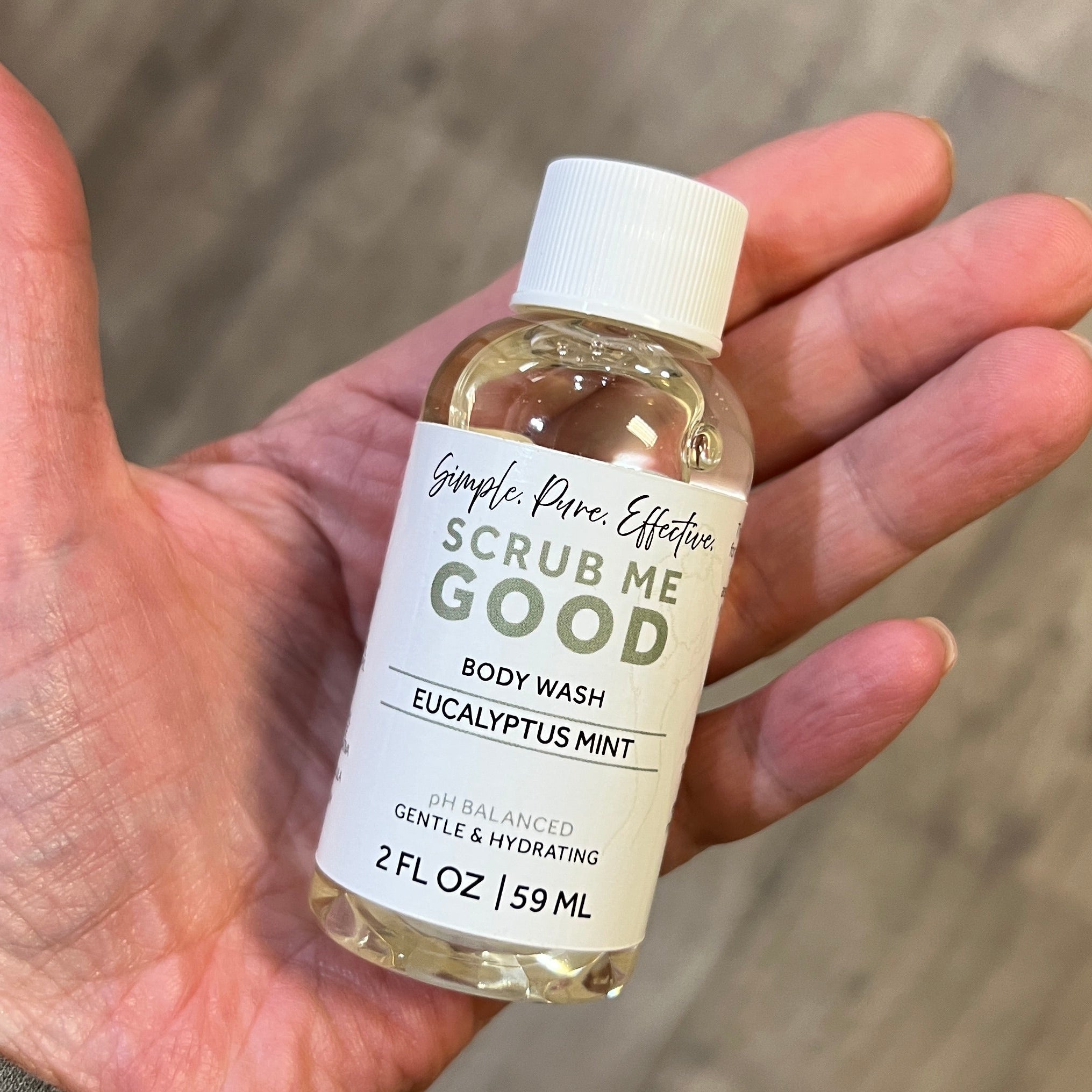 Hand holding a small bottle of body wash labeled 'Scrub Me Good' on a wooden floor.